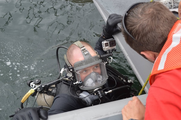 A man in scuba gear is in water looking up at another man who is talking to him from a platform above him.