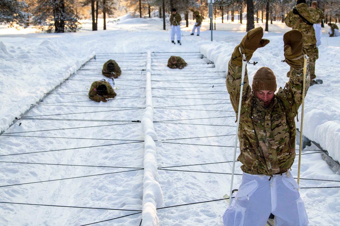 Soldiers low-crawl through an obstacle as another kneels in the snow.
