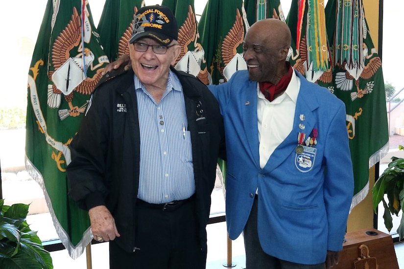 Two men laugh as they pose for a photo with flags behind them.