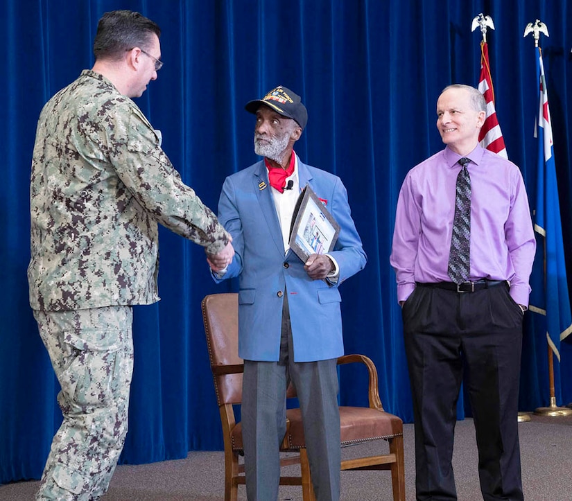 Three men stand on a stage as one shakes the hand of another.