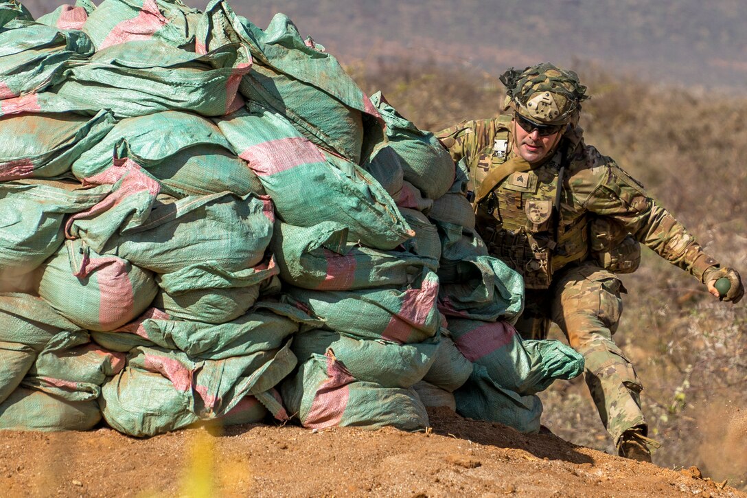 A soldier prepares to throw a grenade while kneeling next to a pile of sandbags.
