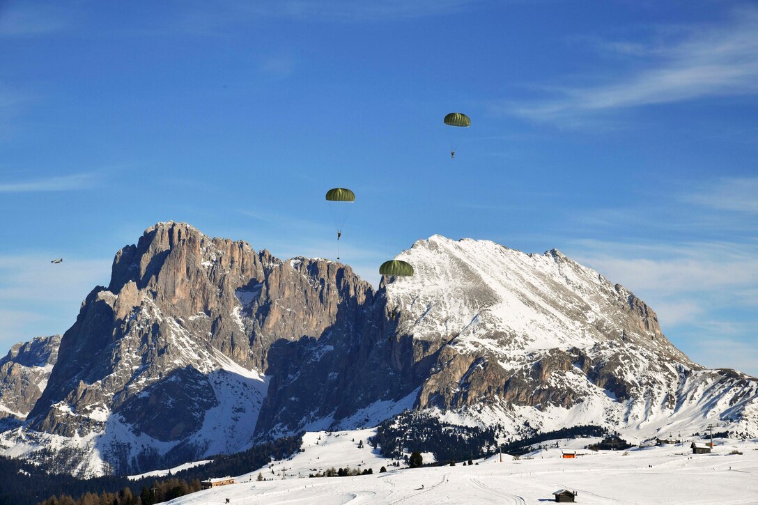 U.S. and Italian soldiers free fall with parachutes over a snow mountain as an aircraft flies nearby.