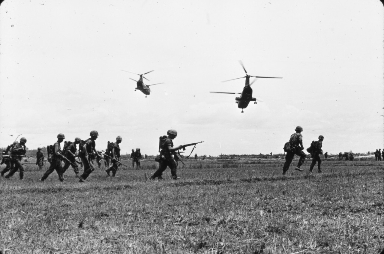 Several men with guns run through a field as two helicopters hover overhead.