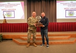 a white male in a U.S. Army camo military uniform hands a large globe-shaped trophy to a white male in black sweater and blue jeans in front of a small auditorium stage.