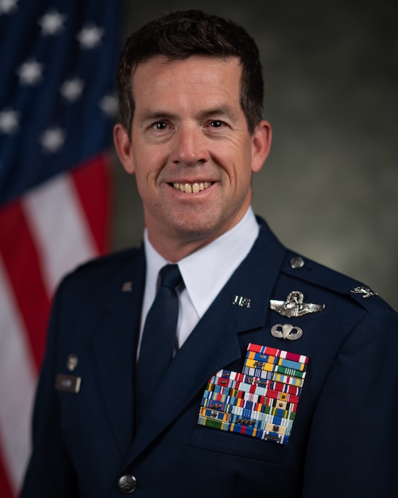 An official head and shoulders portrait Air Force biography photo depicting Col. Matthew R. Glynn in his service dress uniform with a subdued gray background including a U.S. flag hanging behind the right shoulder.