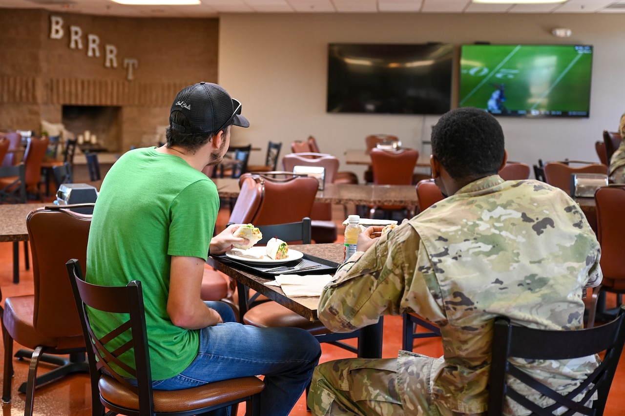 Two young men watch TV while eating at a table in a dining facility.