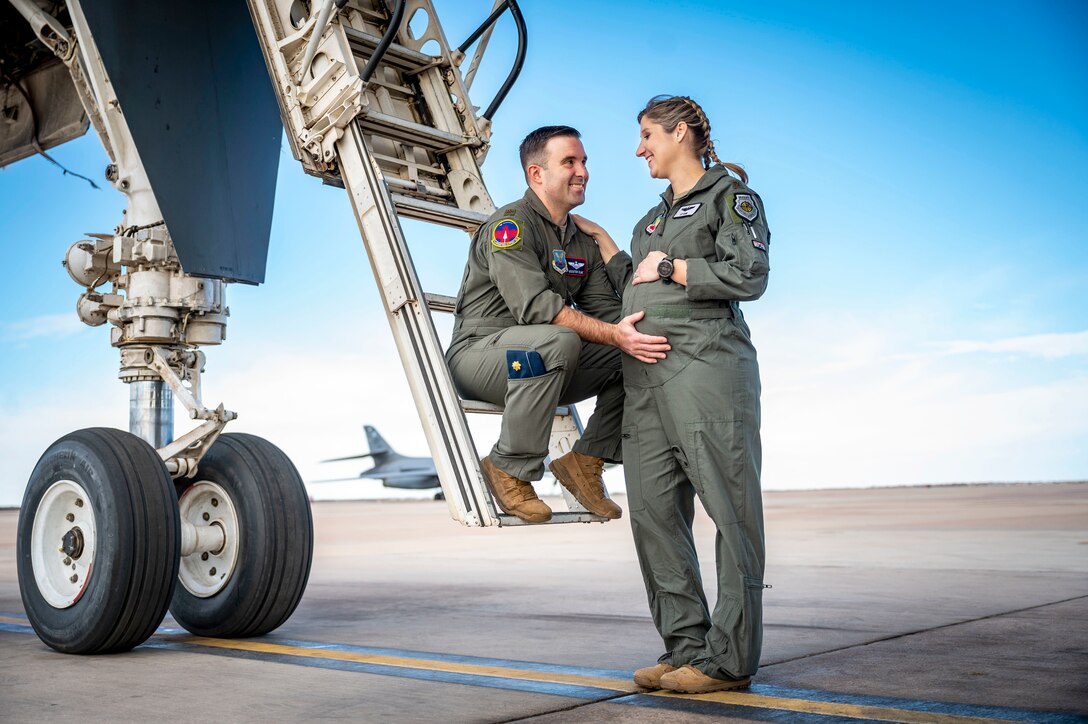 A pregnant pilot stands by an aircraft ladder where her husband sits, with a hand on her belly.