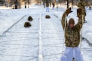 Soldiers low-crawl through an obstacle as another kneels in the snow.