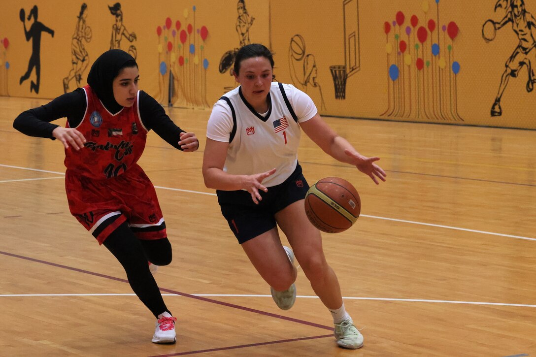 A soldier in a basketball uniform dribbles the ball past another player.