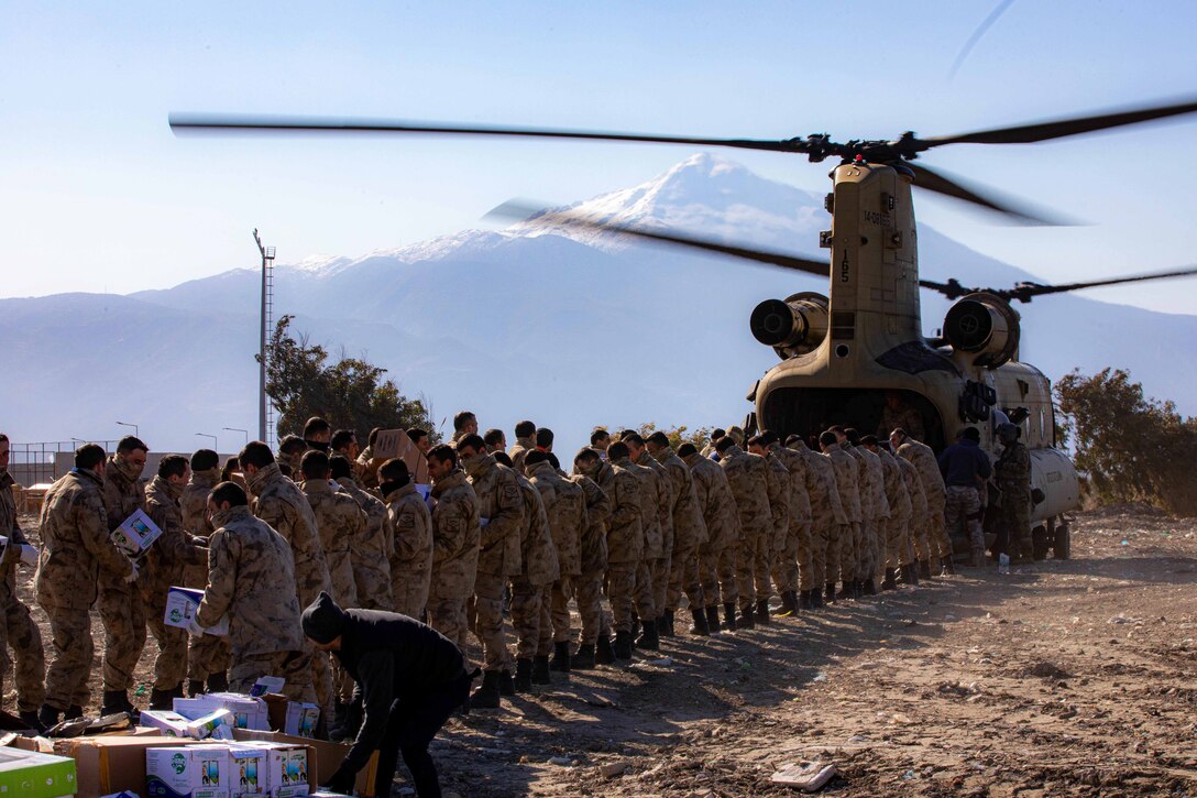 Service members stand side by side in two rows carrying boxes of relief supplies behind a helicopter.