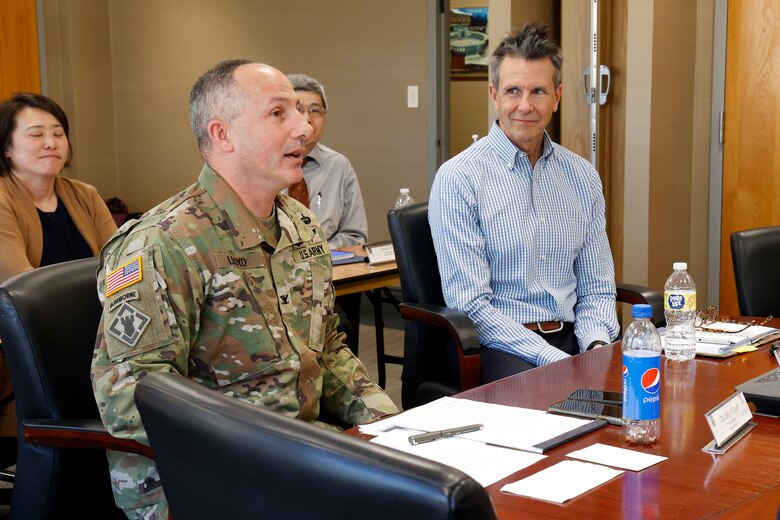 A man in an Army uniform speaks at a meeting while another man seated next to him listens.