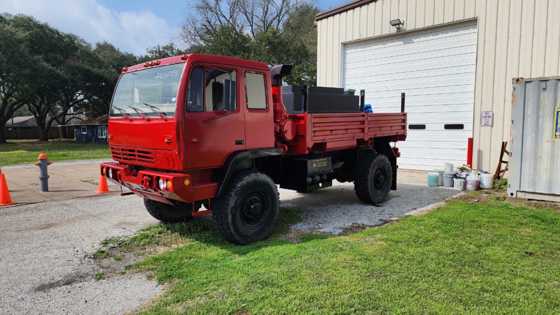 A former military cargo truck painted red sits outside a tan building. The truck has no military markings but does have a water tank on its bed used to take water to fires.