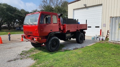 A former military cargo truck painted red sits outside a tan building. The truck has no military markings but does have a water tank on its bed used to take water to fires.