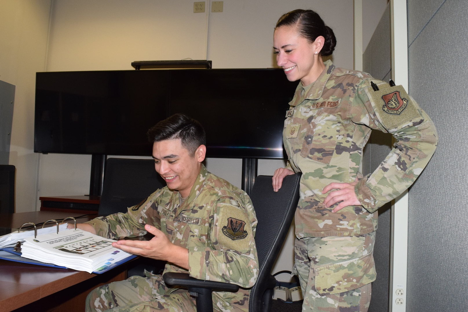 photo of two U.S. Air Force Airmen in uniform, one sitting, one standing in an office looking at a binder