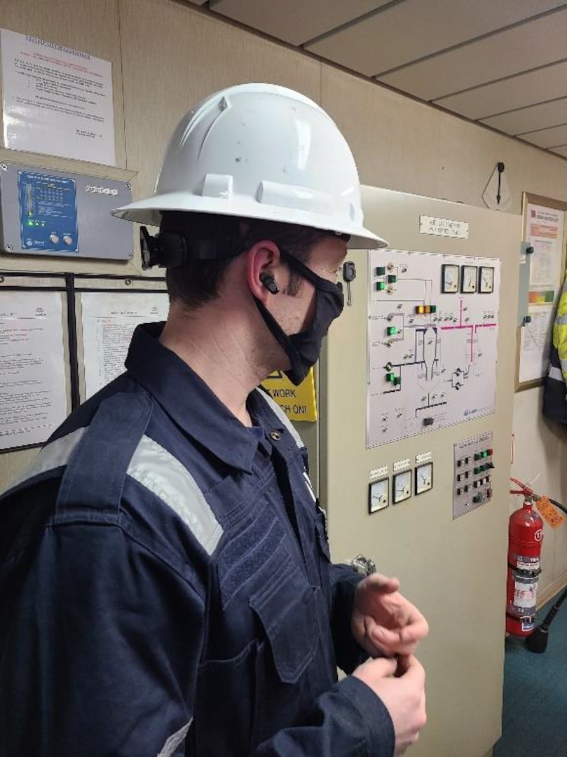 Petty Officer 1st Class Bradley Martin, a marine science technician, talks with an engineer while wearing noise cancelling earbuds in the engine control room during a Port State Control Exam in Boston, in January 2022. The Coast Guard is testing OTTO electronic hearing protection earbuds to provide enhanced hearing protection in high-decibel environments. (Coast Guard photo)