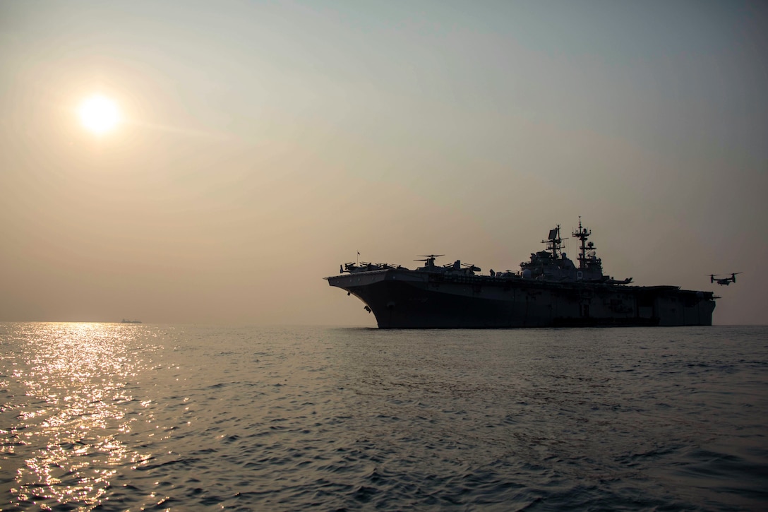 An aircraft prepares to land on a ship at sea under a sunlit sky.