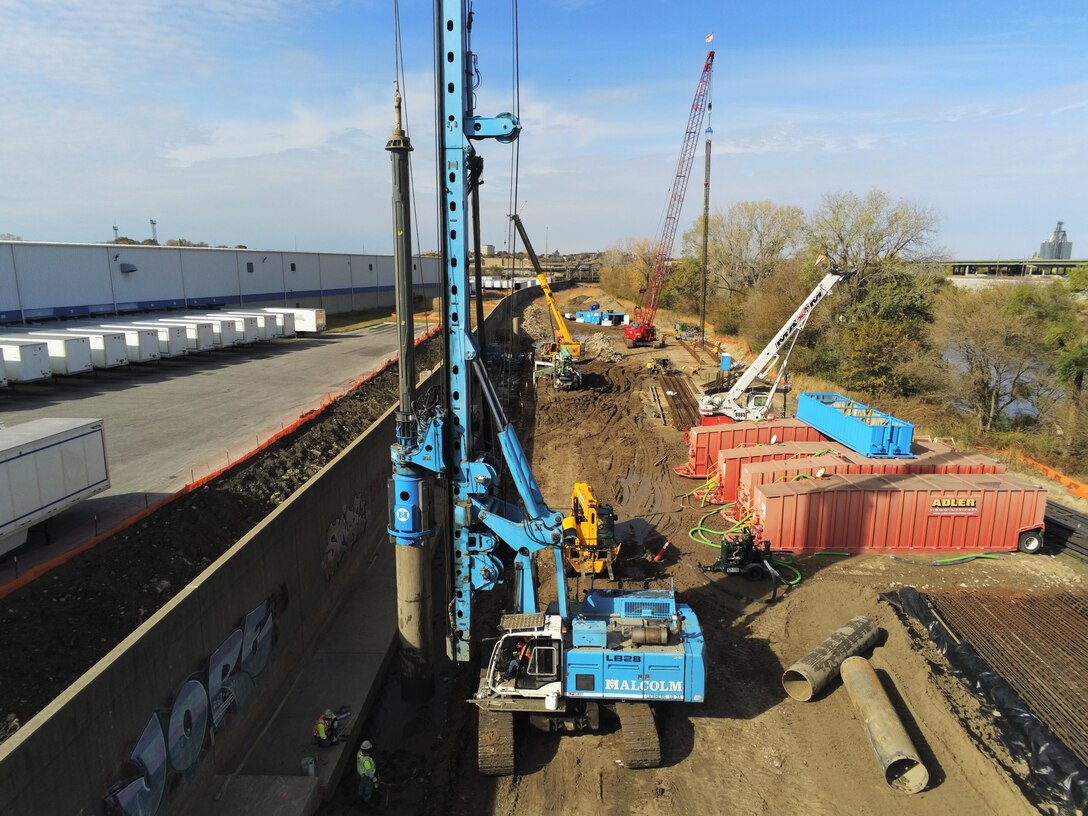 Heavy equipment works on a floodwall with trees in the background.