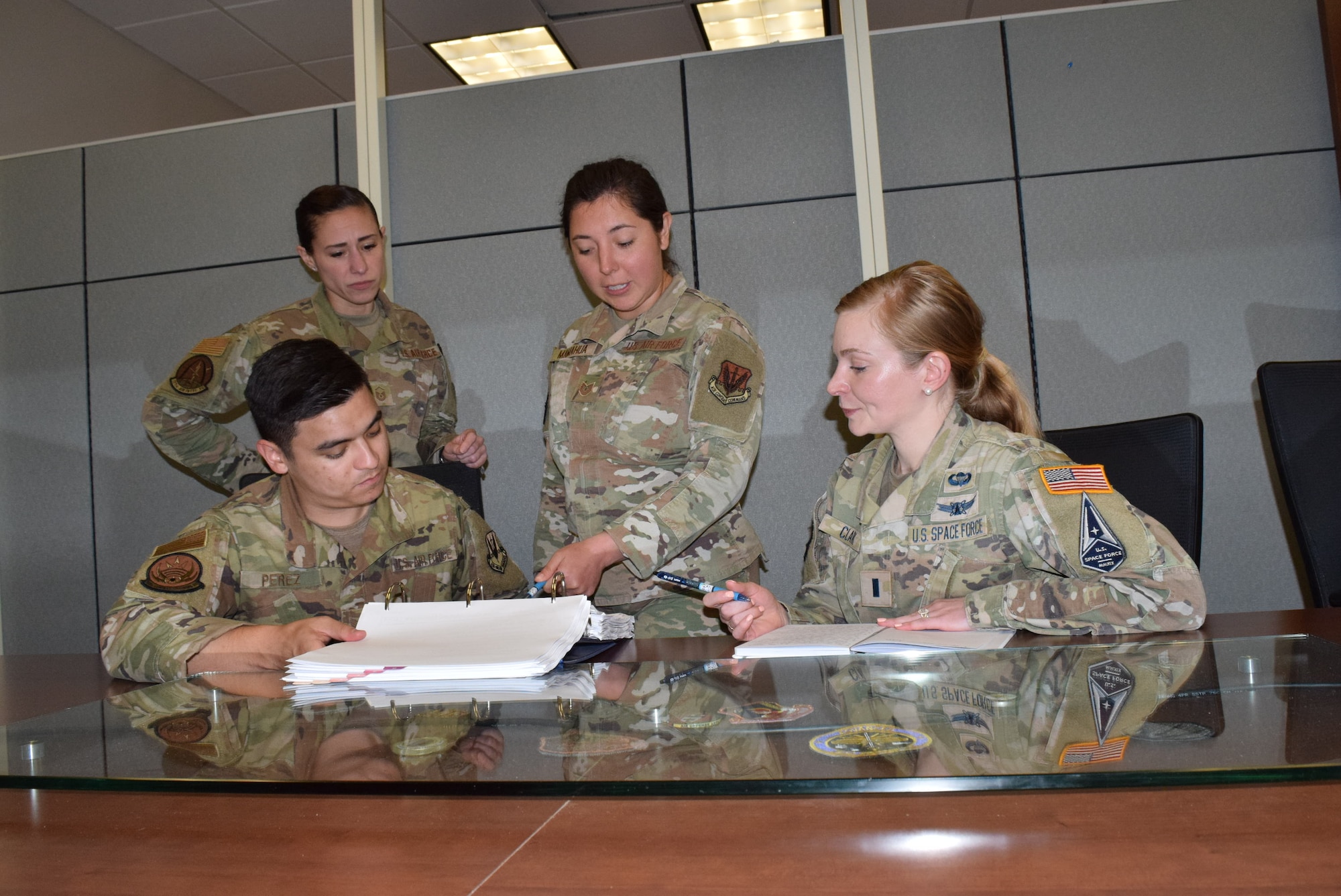 photo of three U.S. Air Force Airmen, one sitting, two standing, and one U.S. Space Force Guardian, sitting reviewing information in a binder.