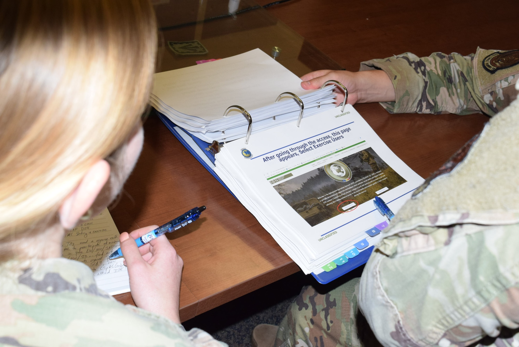 Over the shoulder photo of two U.S. Air Force Airmen in uniform looking at a blue binder.