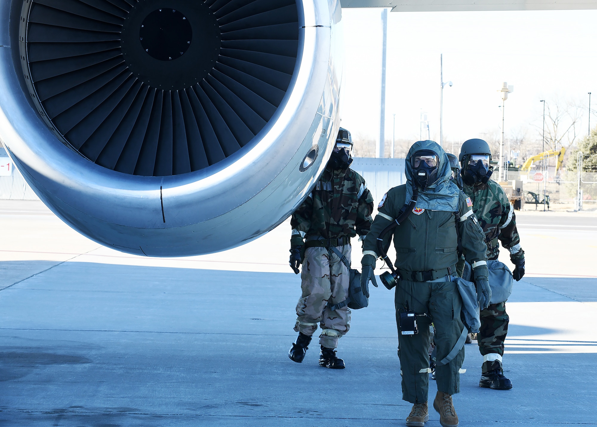 Three Airmen in MOPP gear walk by an engine aircraft