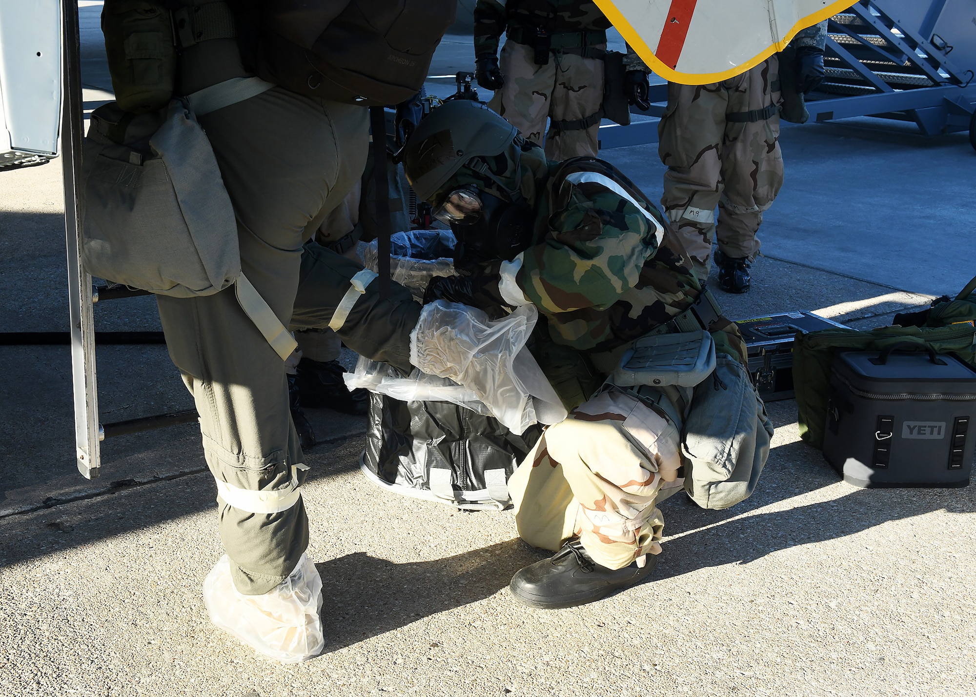 Airman in MOPP gear removes boot covering on another Airmen