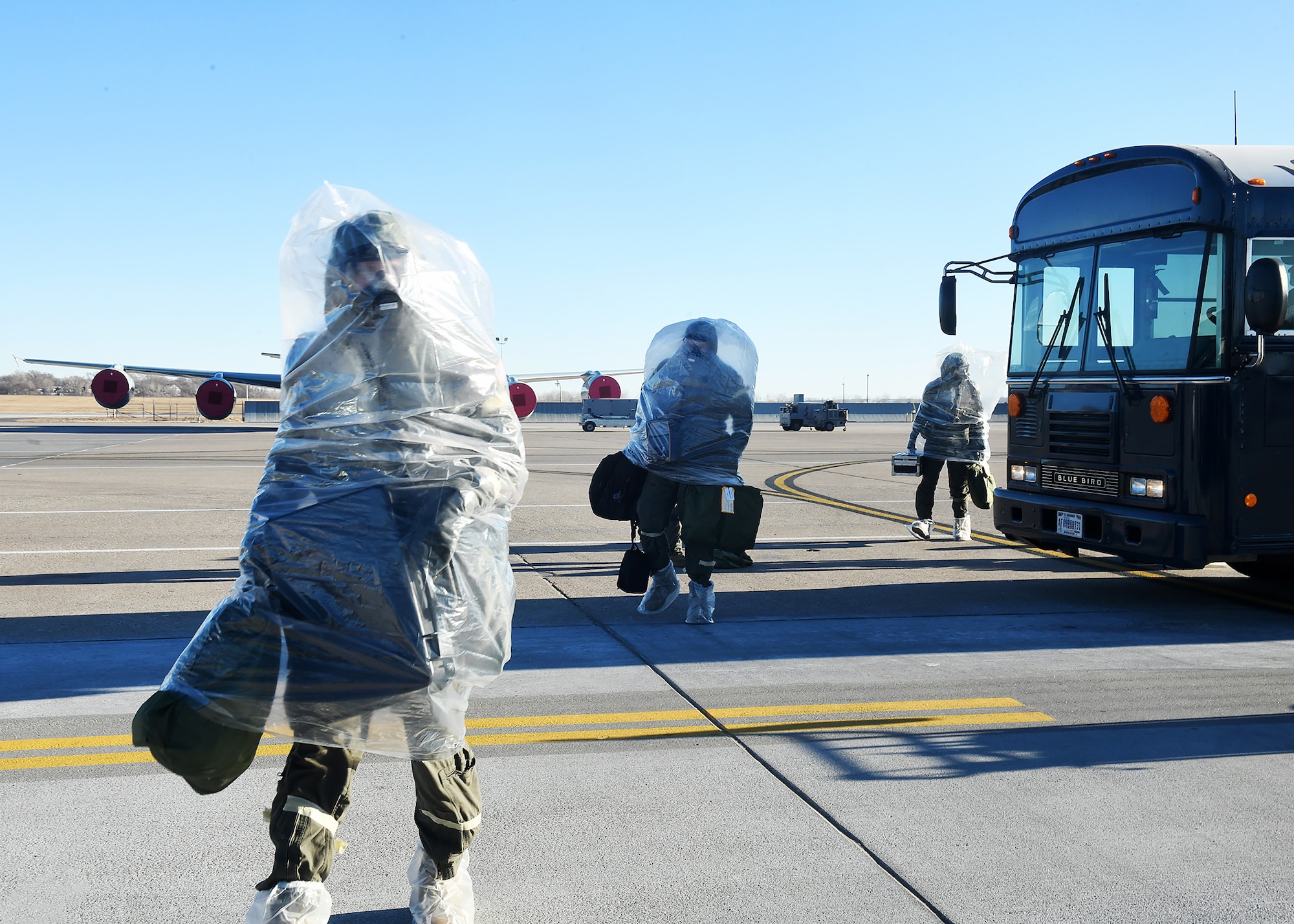 Three Airmen in MOPP gear walking in front of a bus and an aircraft