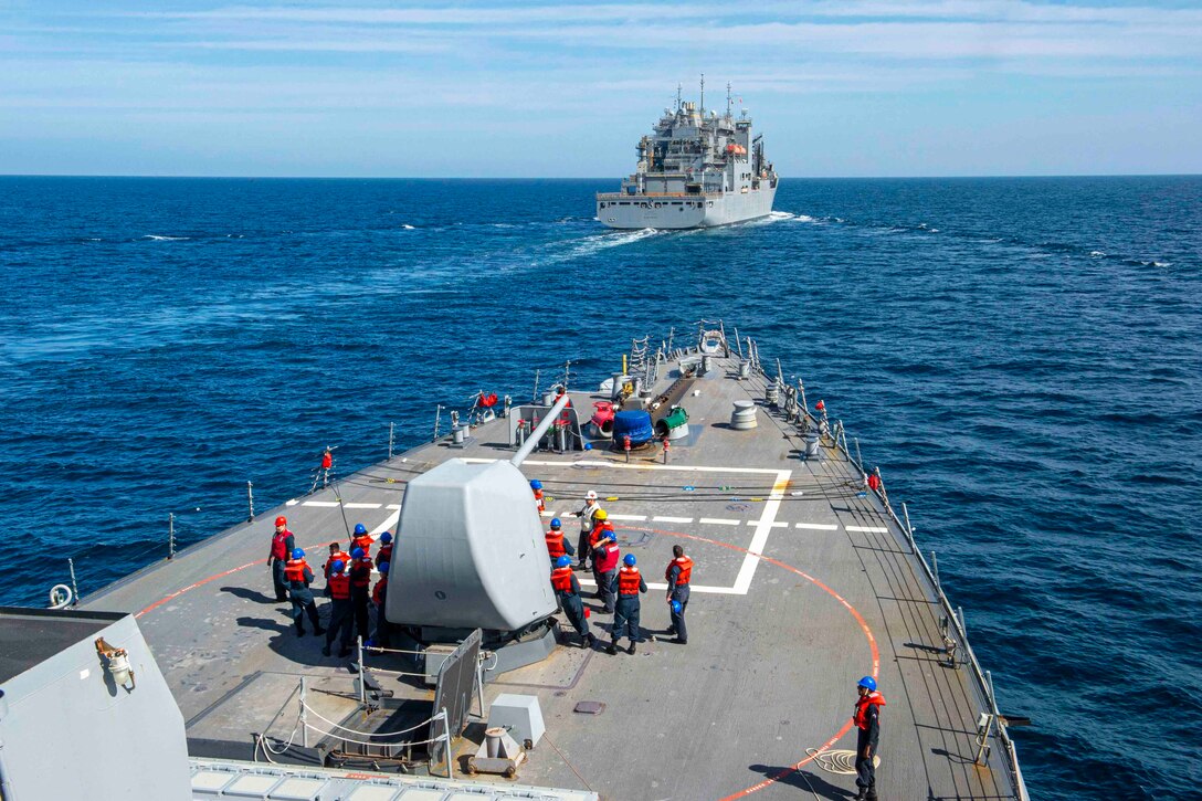 Sailors stand on the deck of a ship as it approaches another ship.