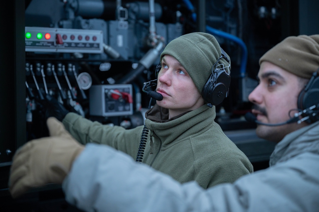 Two service members wearing headsets look at a control panel.