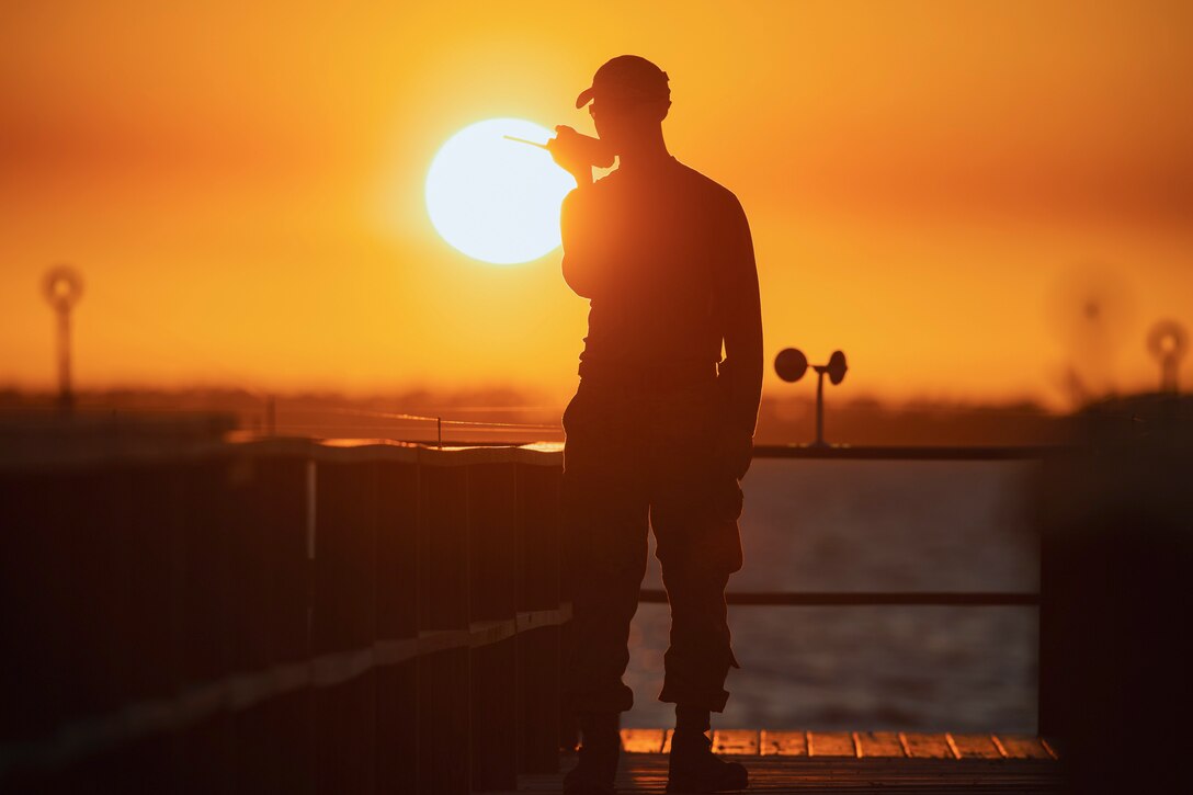 An airman talks into a hand-held communicatons device with an orange sunlit sky backdrop.