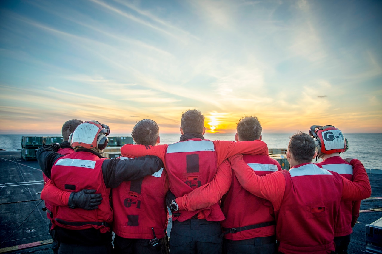 Seven sailors put their arms around each other as they watch the sunset aboard a ship at sea.