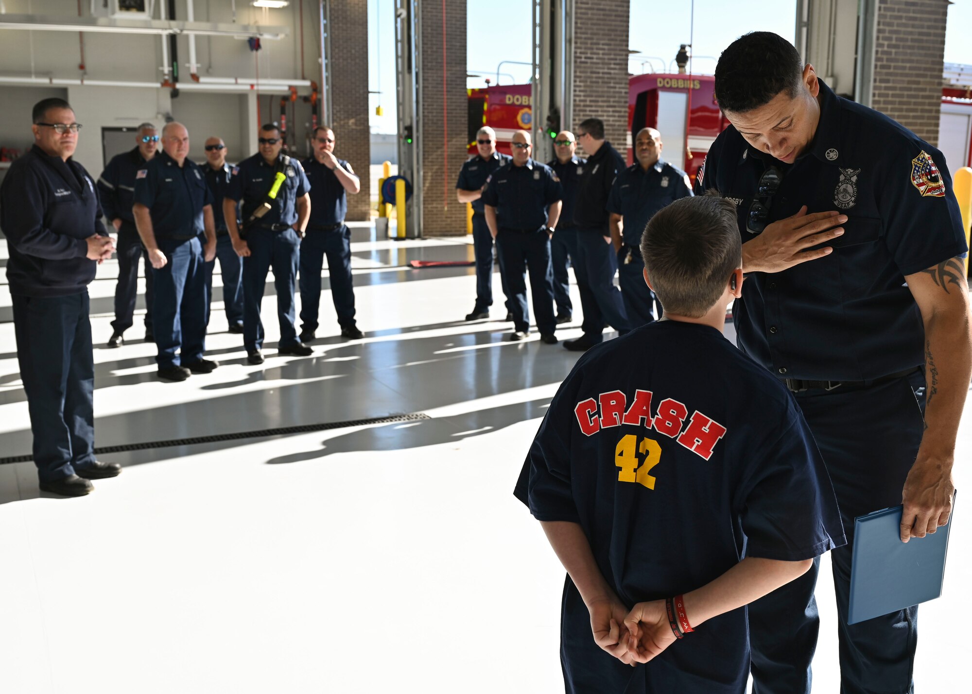 Man in blue firefighters uniform shows a boy his badge inside of a fire station bay.