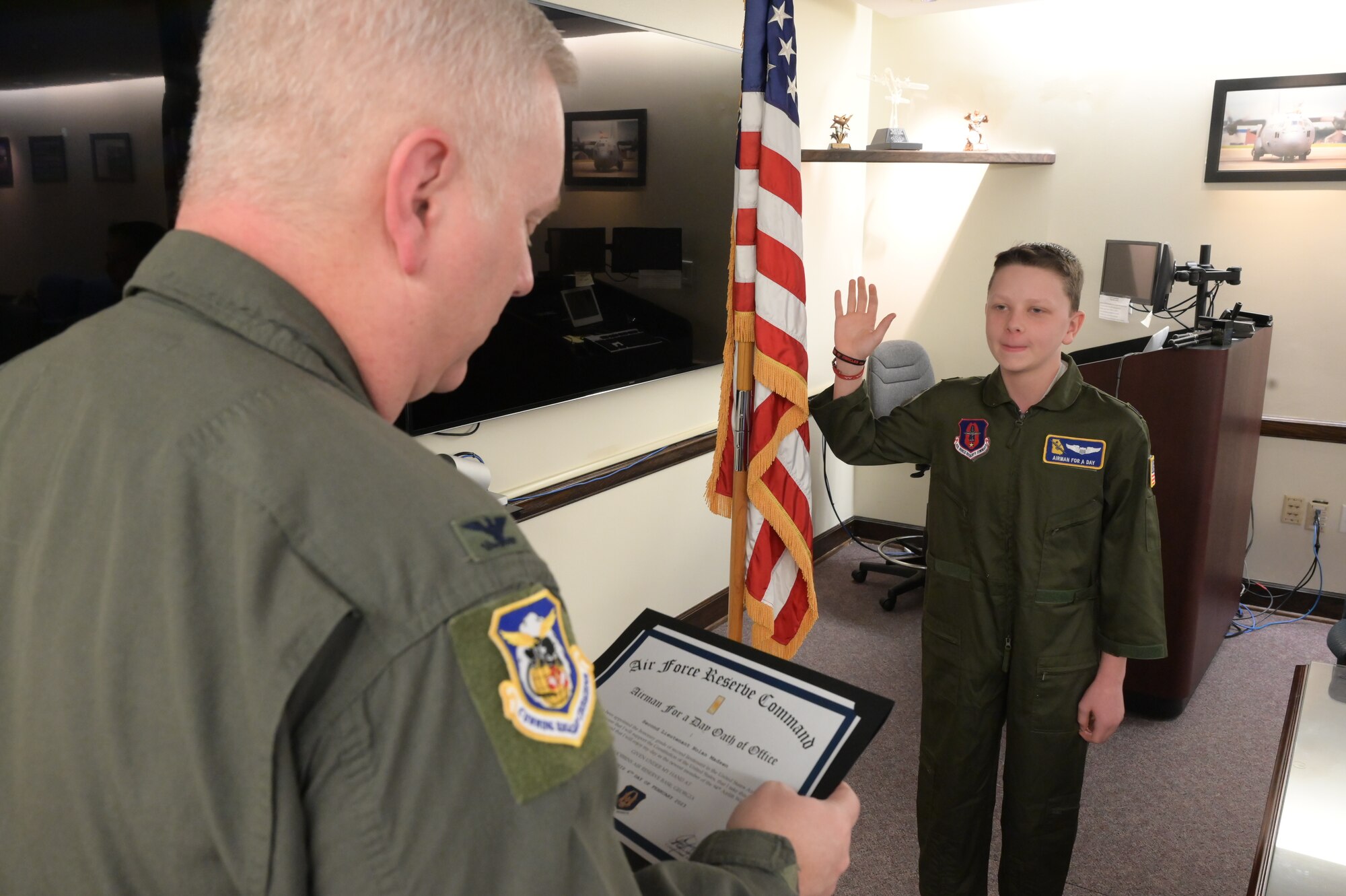 Man in green flightsuit swears a boy in a green flightsuit into "office".