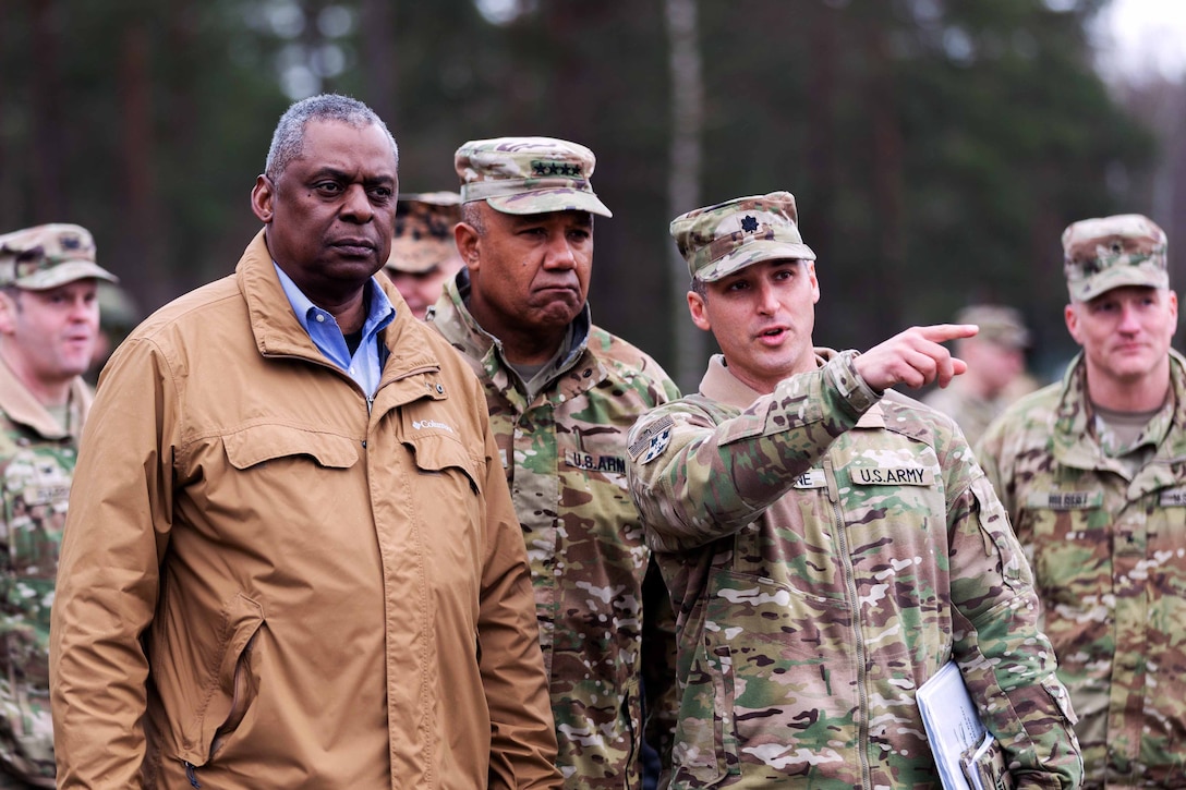 Secretary of Defense Lloyd J. Austin III stands with soldiers as one soldier points to the right.