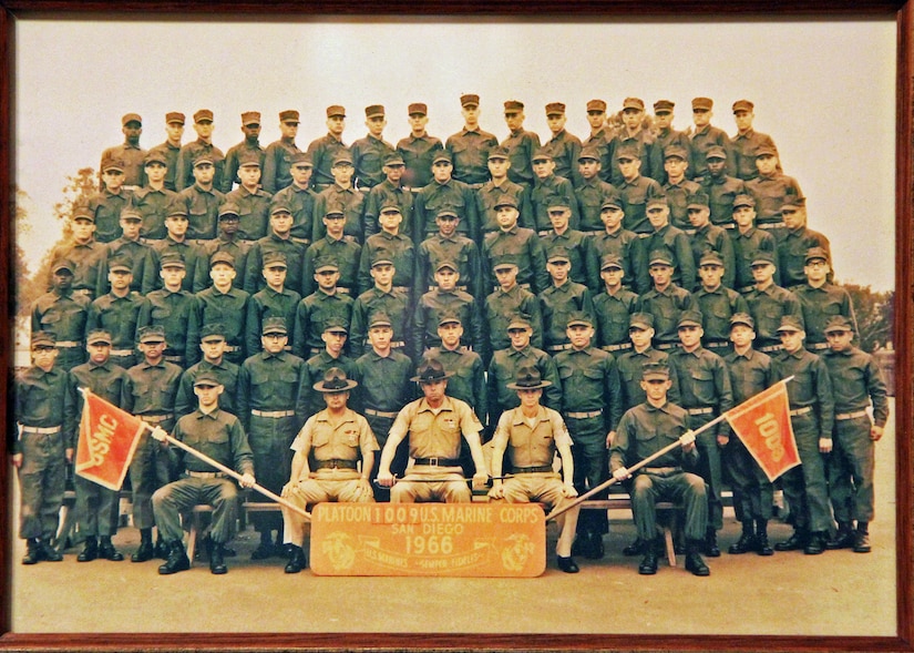 Six tiered rows of men pose for a photo in front of a plaque and two flags.