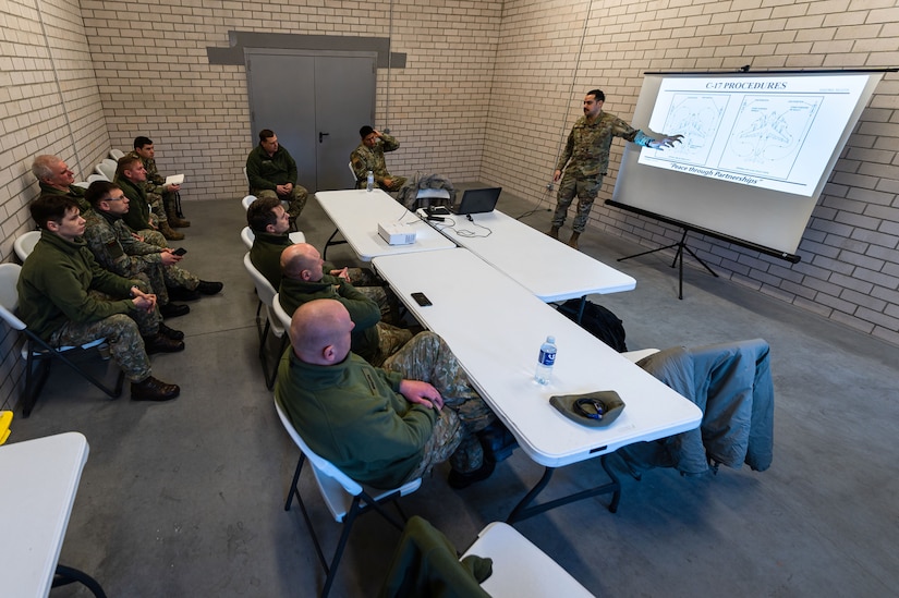 An airman stands and points at text on a large screen in front of a class of fellow troops.