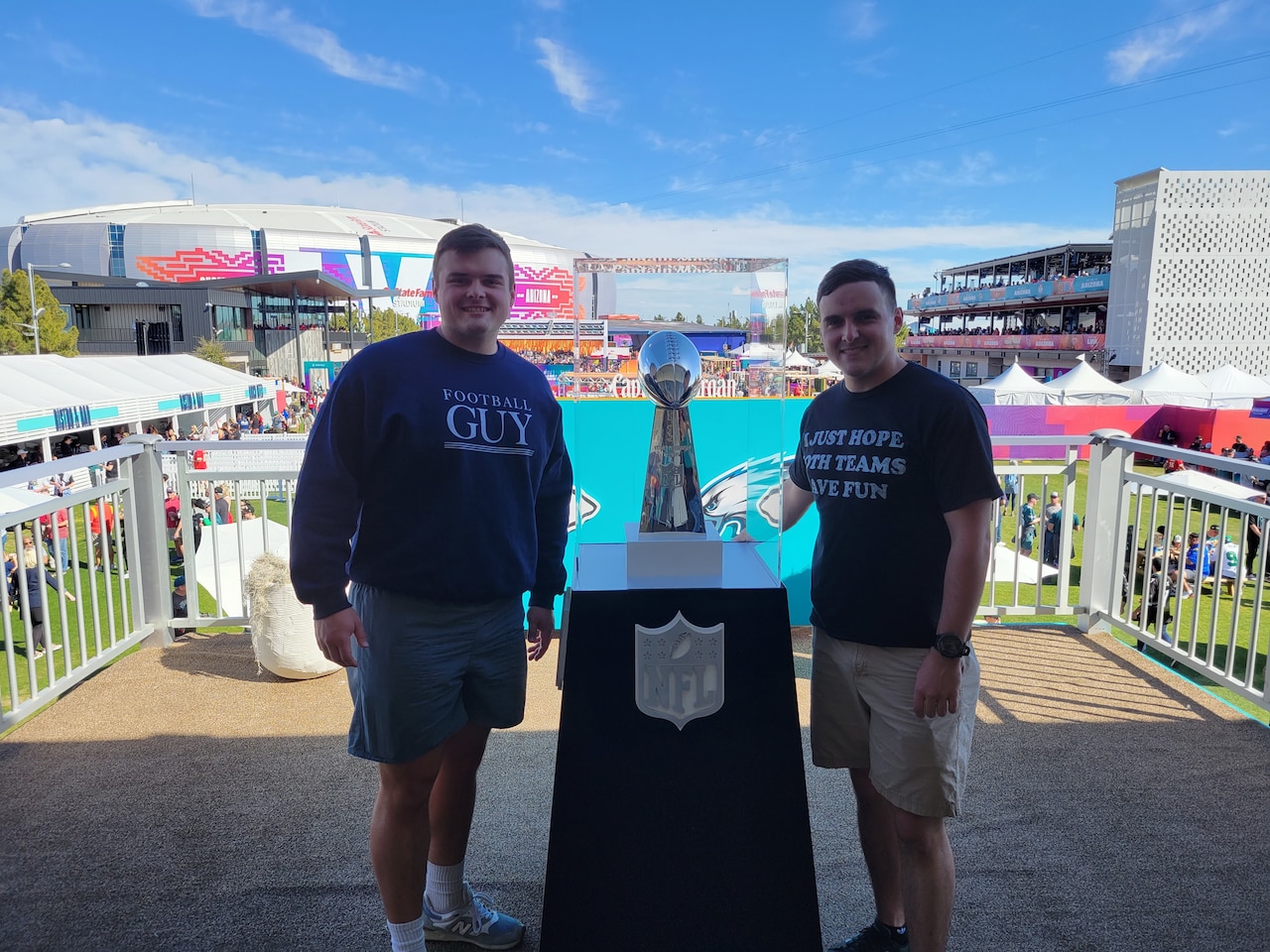 Two men stand beside a trophy in a glass case. A stadium is seen in the background.