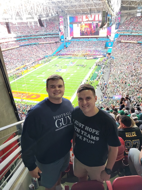 Two men poses for a photo high up in a stadium with a football field in the background.