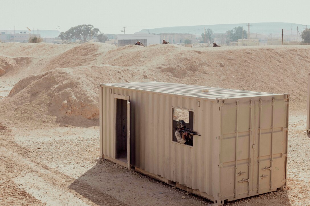 A Marine aims a weapon from a container in a desert-like area as fellow Marine hide behind sand dunes.