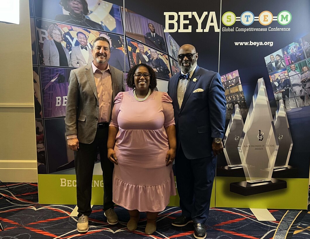 Three adults stand in front of a banner, two men and one woman, and pose for a picture.