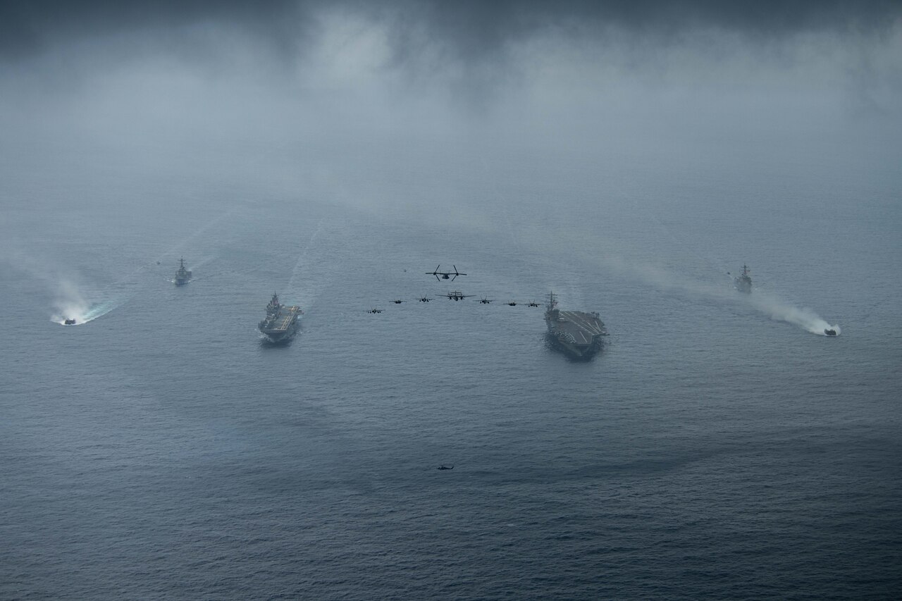 Ships and aircraft move in formation in and above a body of water.