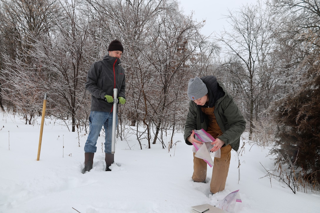 A man with a long metal pole stands next to a woman who is collecting plastic bags. They are both dressed in winter gear and are outside in the snow.