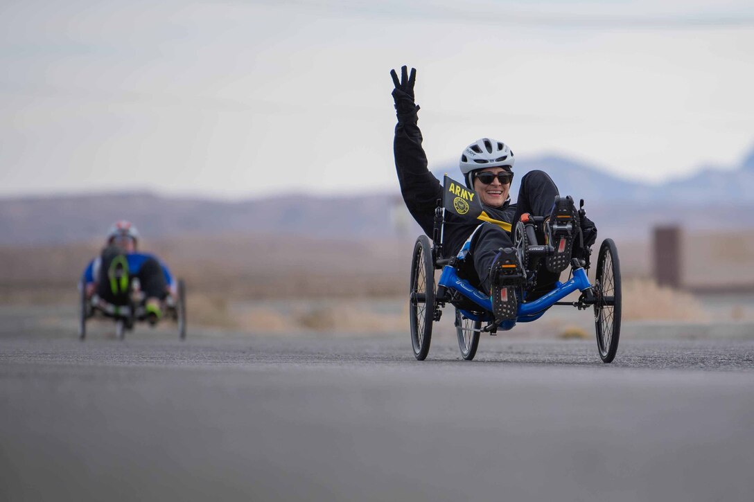 A cyclist raises her arm and displays three fingers as she rides on a road.