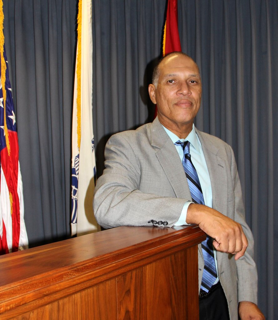 A man leans on a podium in front of three flags.