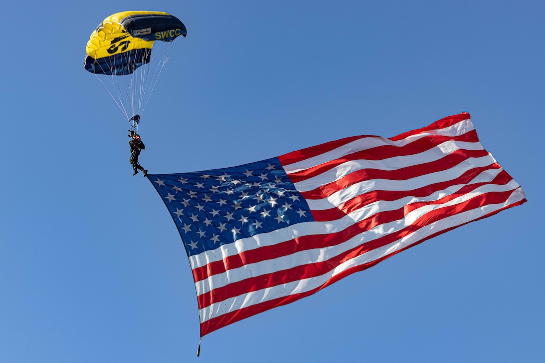 A parachutist with a large U.S. flag attached to his body descends to the ground against a blue sky.