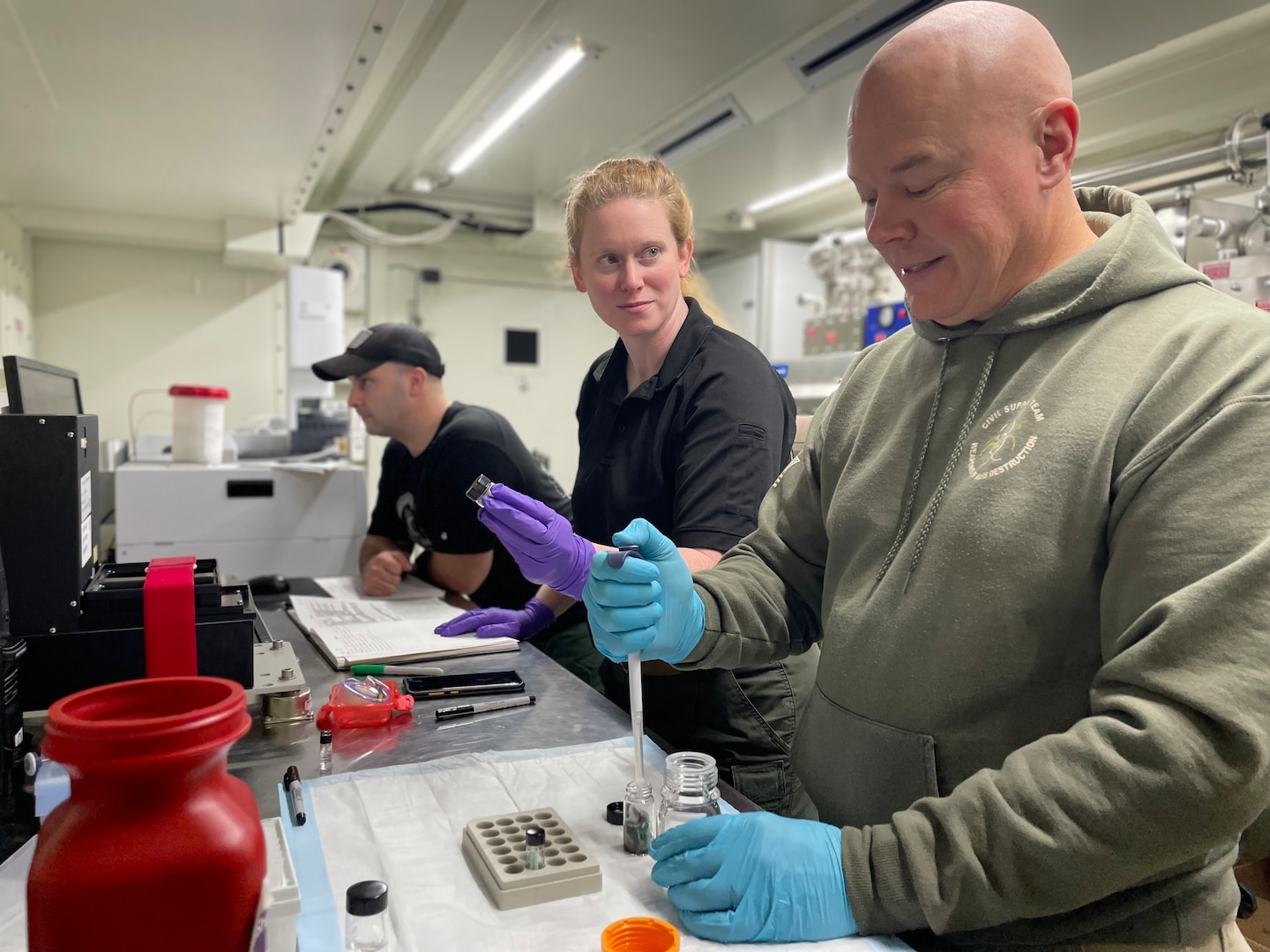 Man wearing olive green sweatshirt and blue latex gloves drops a liquid from dropper into beeker in a small mobile lab.