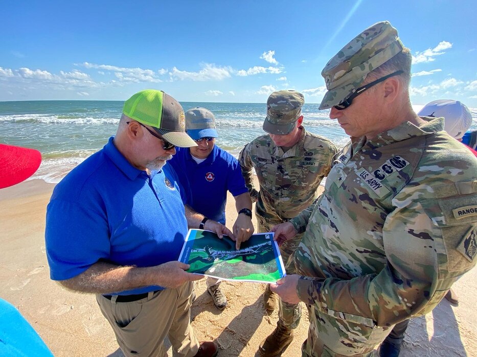 An Army Brig. Gen. and Col. in camouflage uniform review a map with 2 men in blue shirts standing on a beach with the ocean in the the background. 