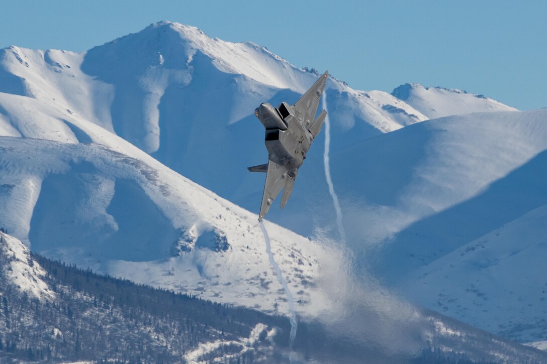 A military aircraft is shown flying sideways with snowy mountains behind it.