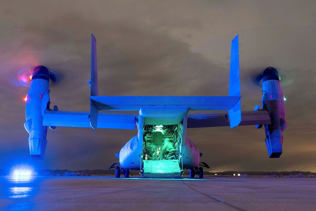 Marines kneel in an open aircraft parked on a tarmac illuminated by colorful lights.