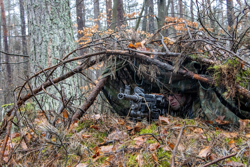 A soldier with a gun rests in a concealed, prone position.