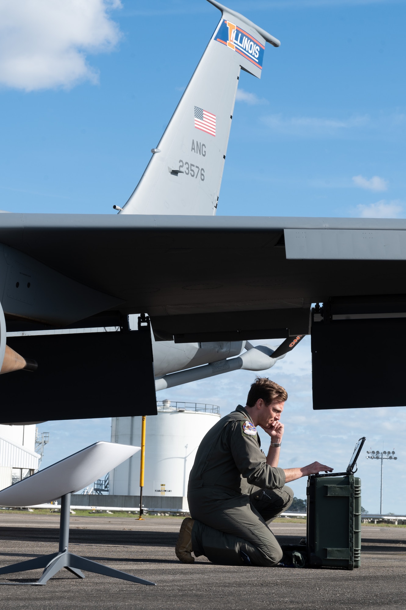 A U.S. Air Force pilot kneels down to type on a laptop that is wirelessly connected to a small satellite dish nearby. A KC-135 tail section is in the background.